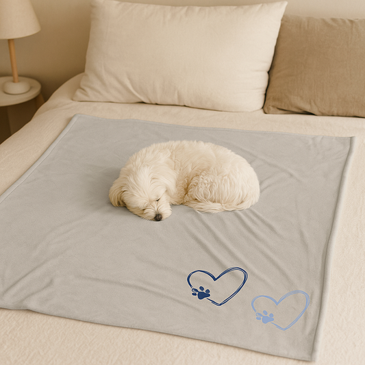 A light gray velveteen blanket with two blue heart and paw print designs, featuring a sleeping Maltese dog on a beige bed.